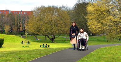 2 women walking down a path in a park. One women is walking and one is in a wheelchair next to her