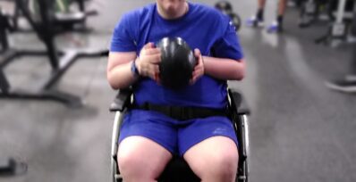 Image shows a young man in a wheelchair at the gym holding a weighted ball