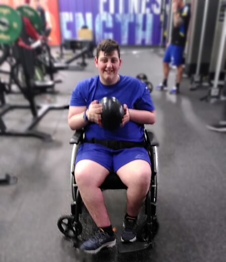 Image shows a young man in a wheelchair at the gym holding a weighted ball